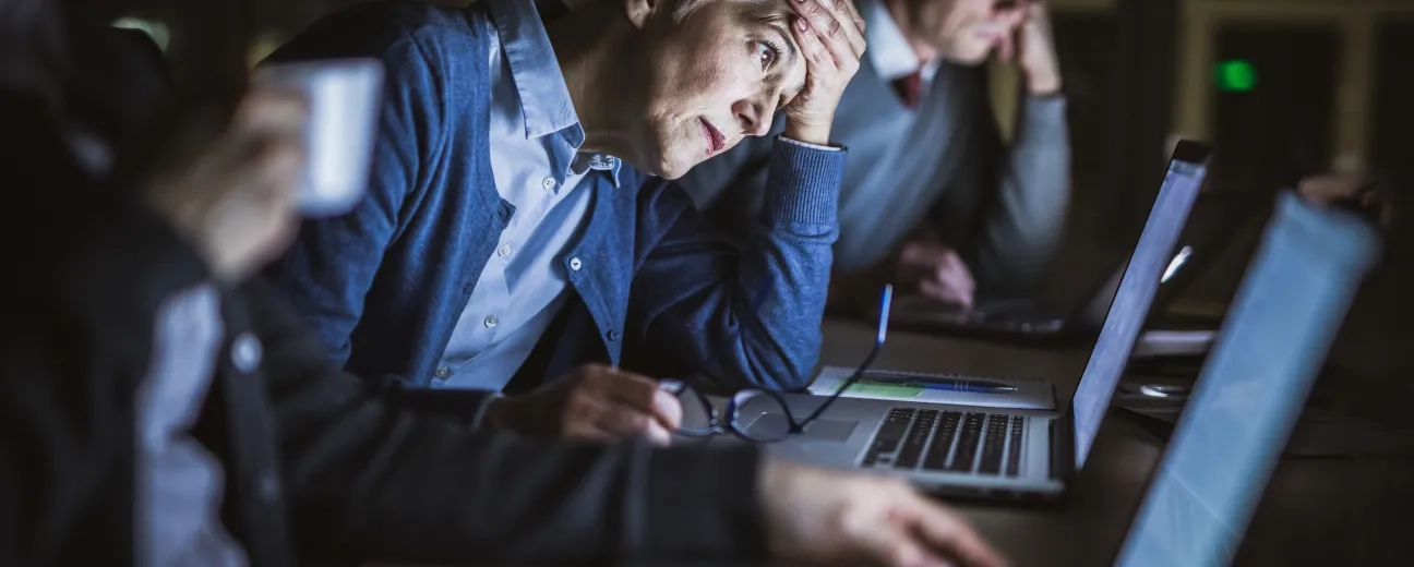 Tired office workers working late at laptops, one person holding their head in frustration.