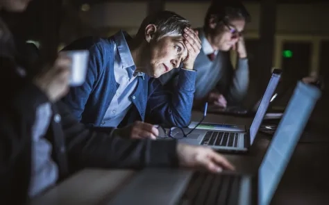 Tired office workers working late at laptops, one person holding their head in frustration.
