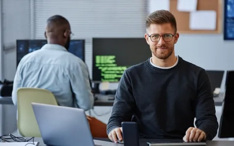 A developer smiling at the camera in an open-plan office, seated at a desk with a laptop, with a colleague working at a monitor displaying green code in the background.