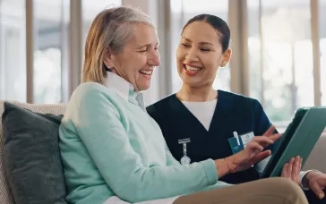 A smiling healthcare professional in a navy uniform assists a happy elderly woman with a tablet while sitting on a couch in a bright, modern setting.