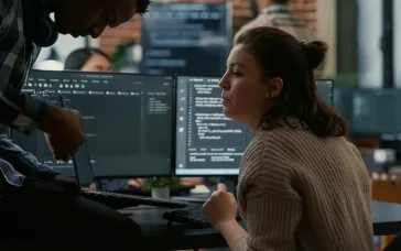 Two coworkers collaborate at a desk in a modern office, reviewing code on dual computer monitors while one person holds a laptop and the other looks on attentively.