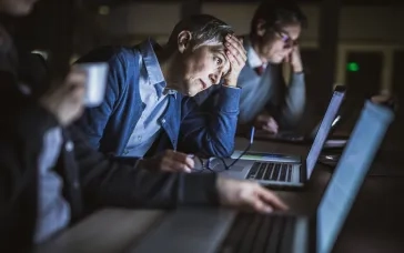 Tired office workers working late at laptops, one person holding their head in frustration.