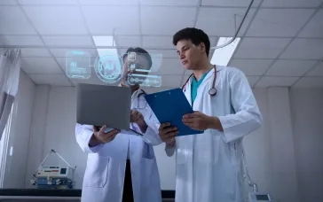 Two healthcare professionals reviewing patient data on a laptop with AI interface overlays in a clinical setting.